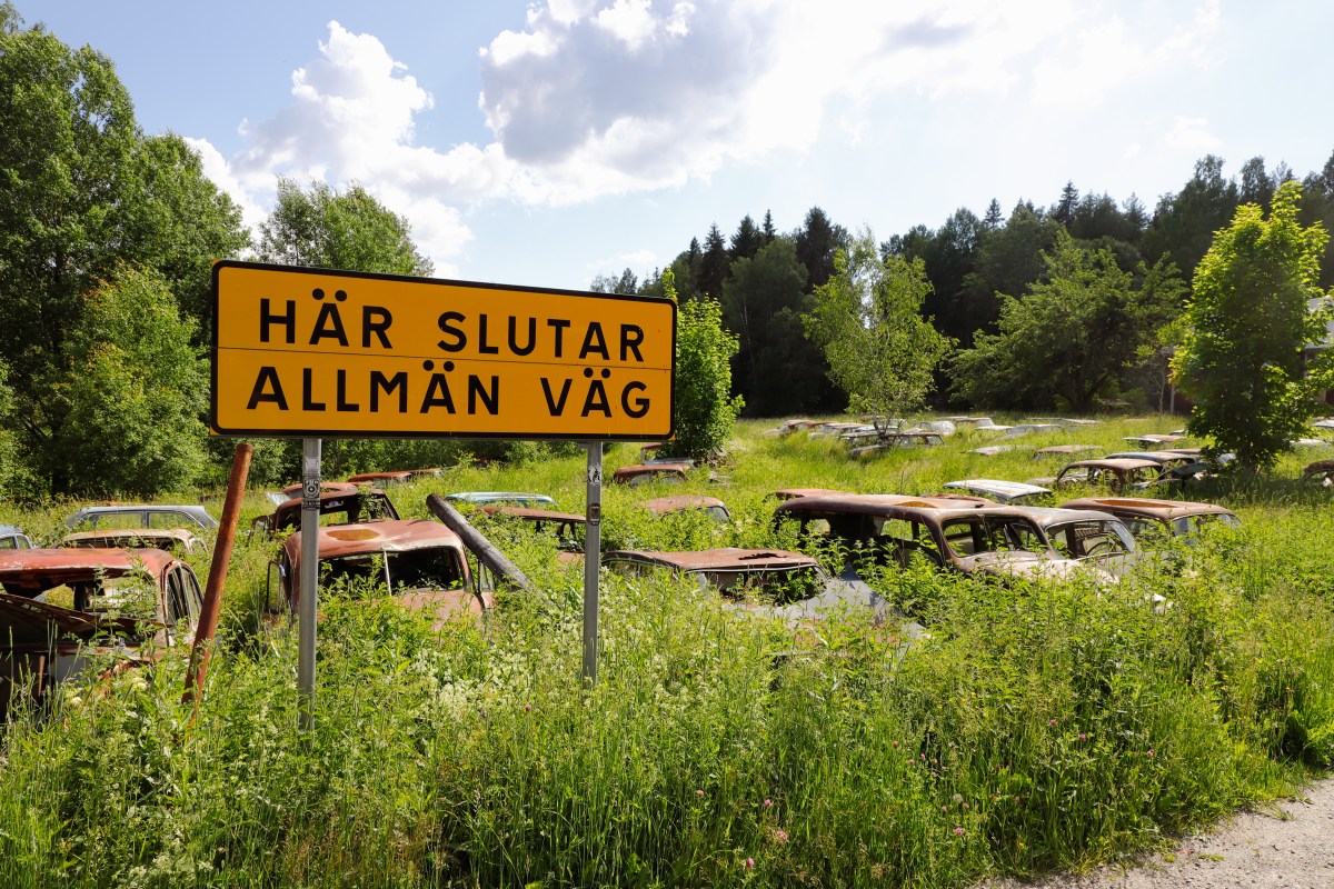 En gul skylt på landsbygd med texten "Hjälp här slutar allmän väg" med flera tomma bilkarosser i bakgrunden.